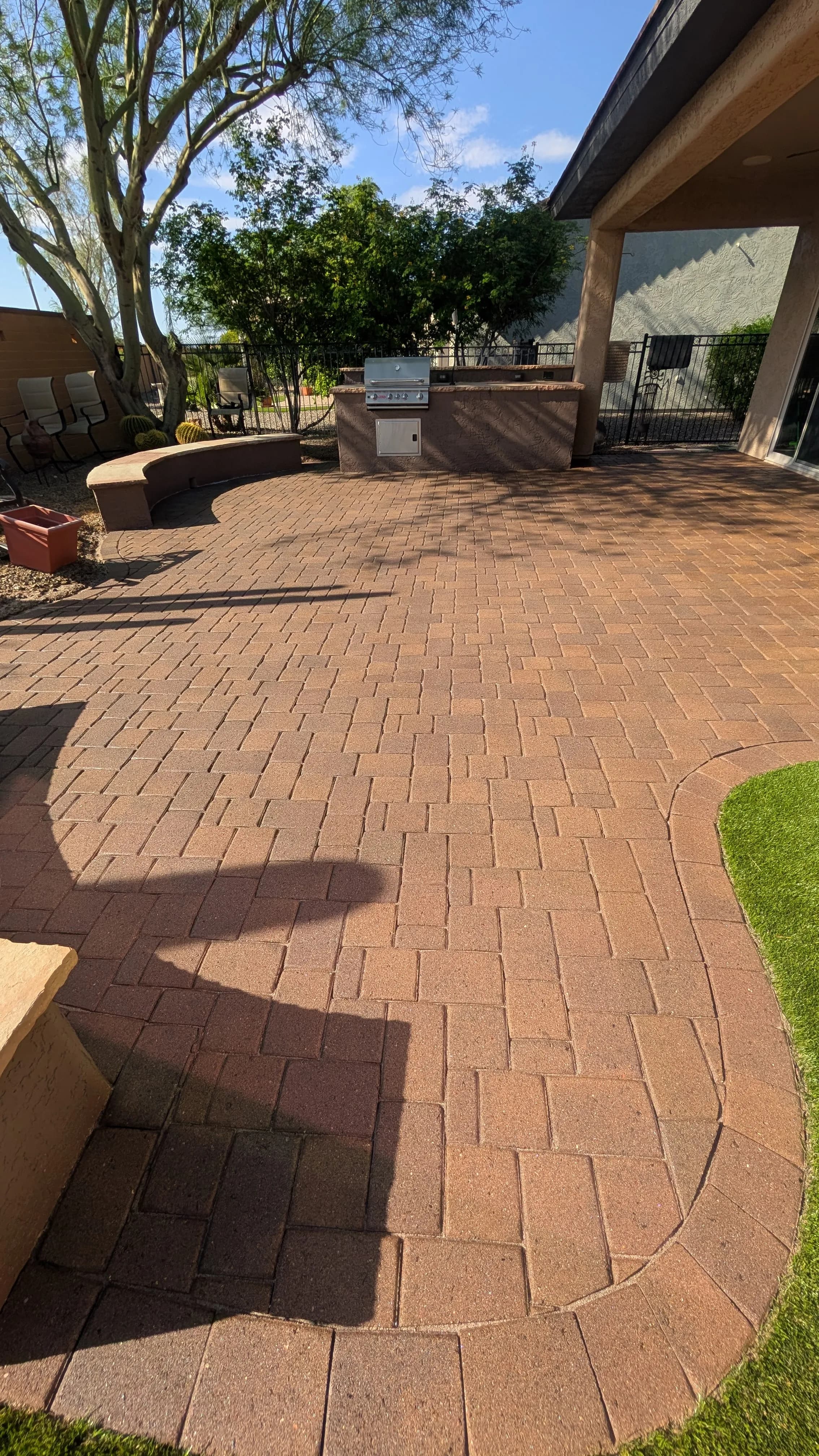 Paved outdoor patio with grill and seating area surrounded by greenery and blue sky.