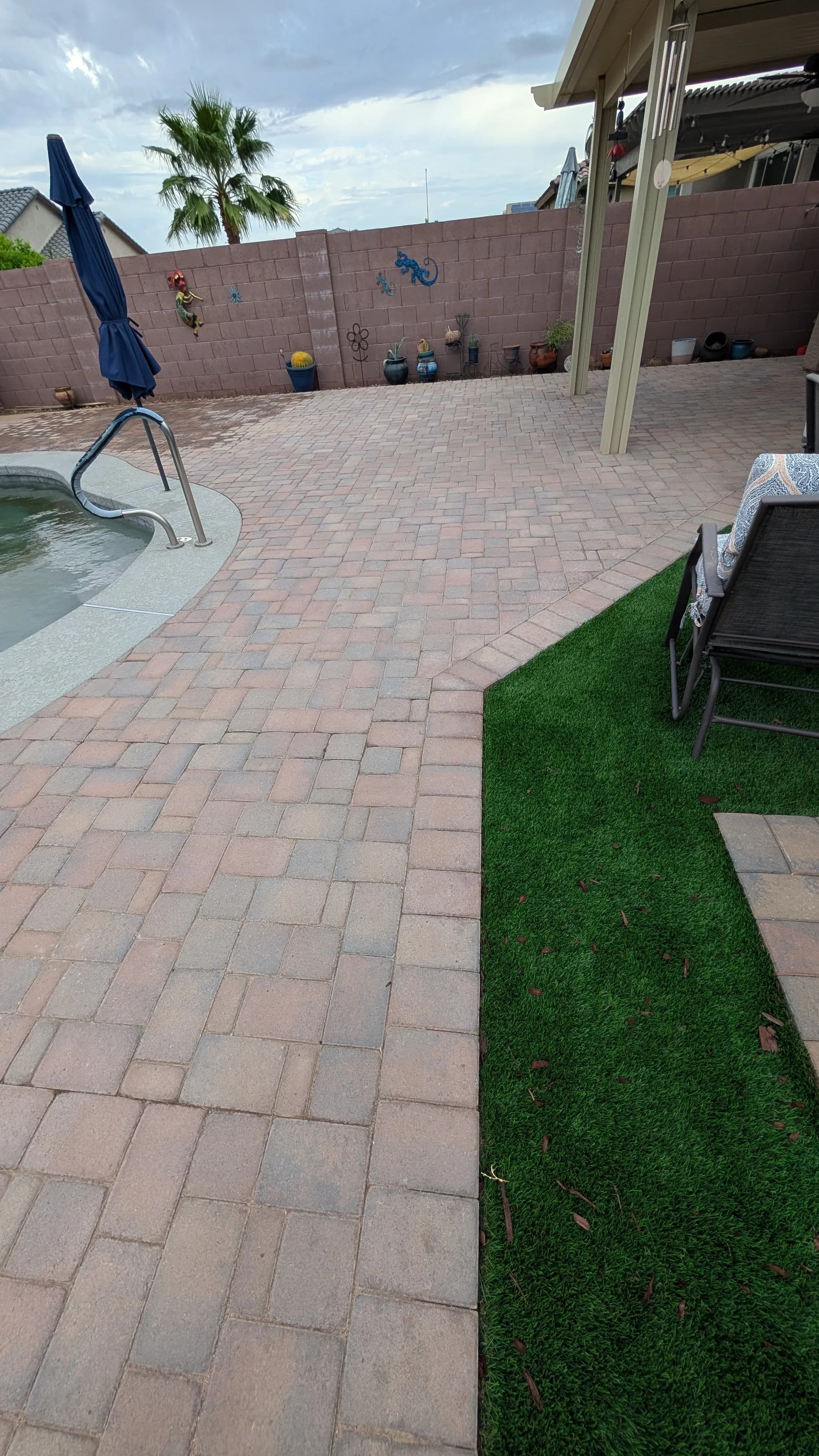 Paved backyard patio with swimming pool, green turf, and wall decor in sunny setting.
