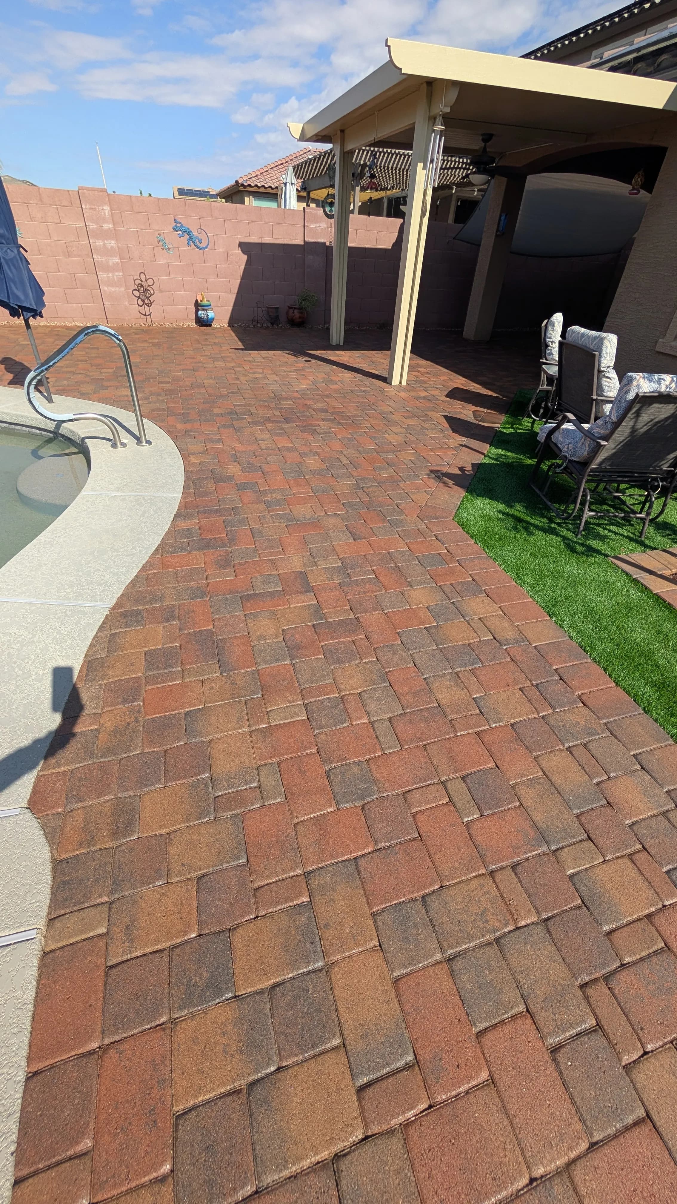 Paved backyard with a swimming pool, grassy area, and covered patio in bright sunlight.