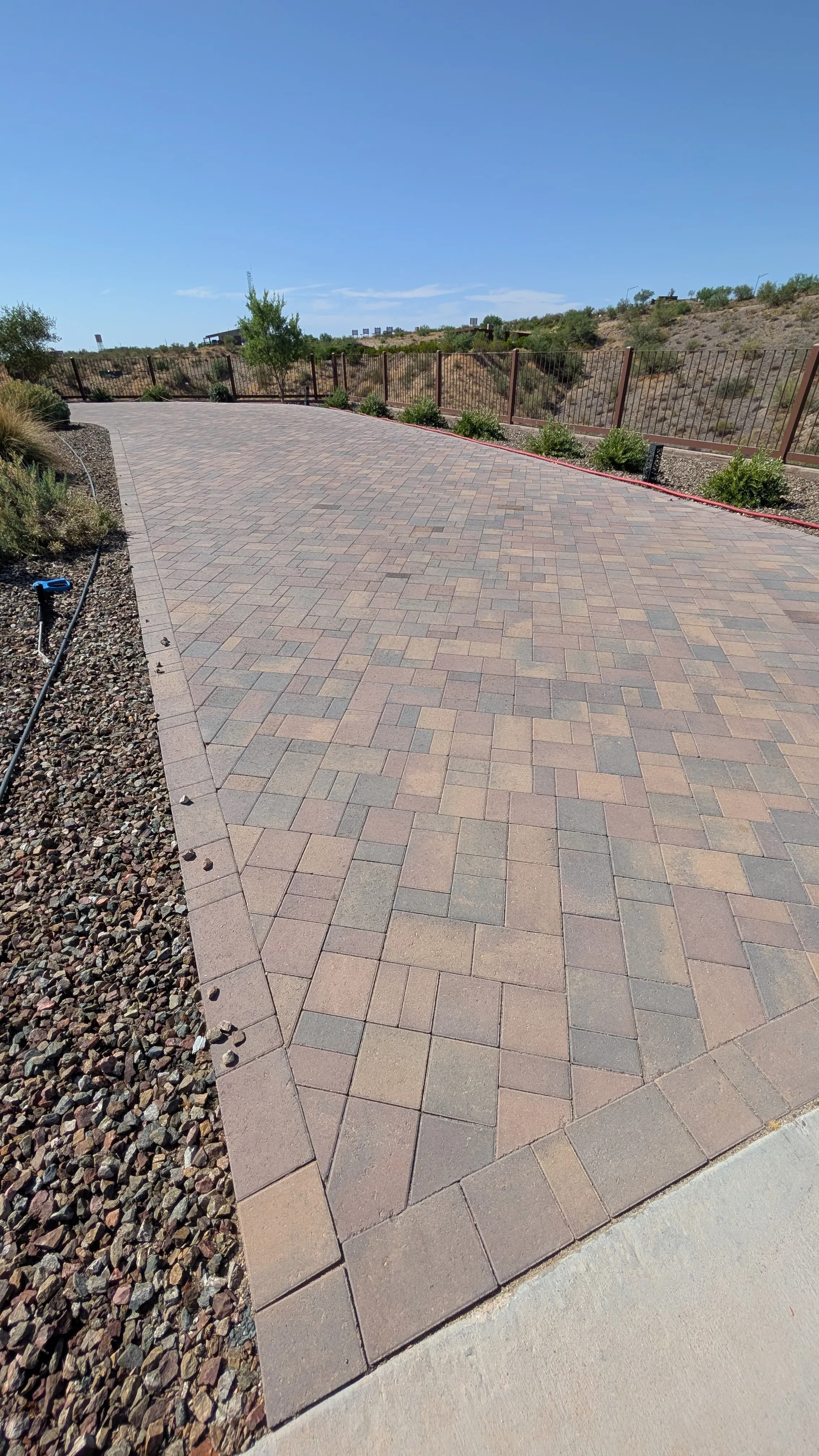Paved driveway with interlocking stones, surrounded by gravel and shrubs under clear blue sky.