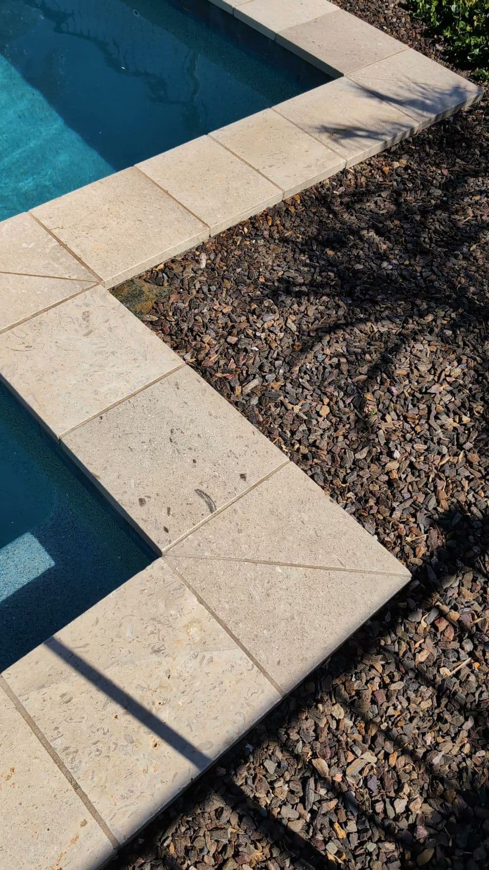 Swimming pool corner with stone coping and pebbled landscaping under sunlight.