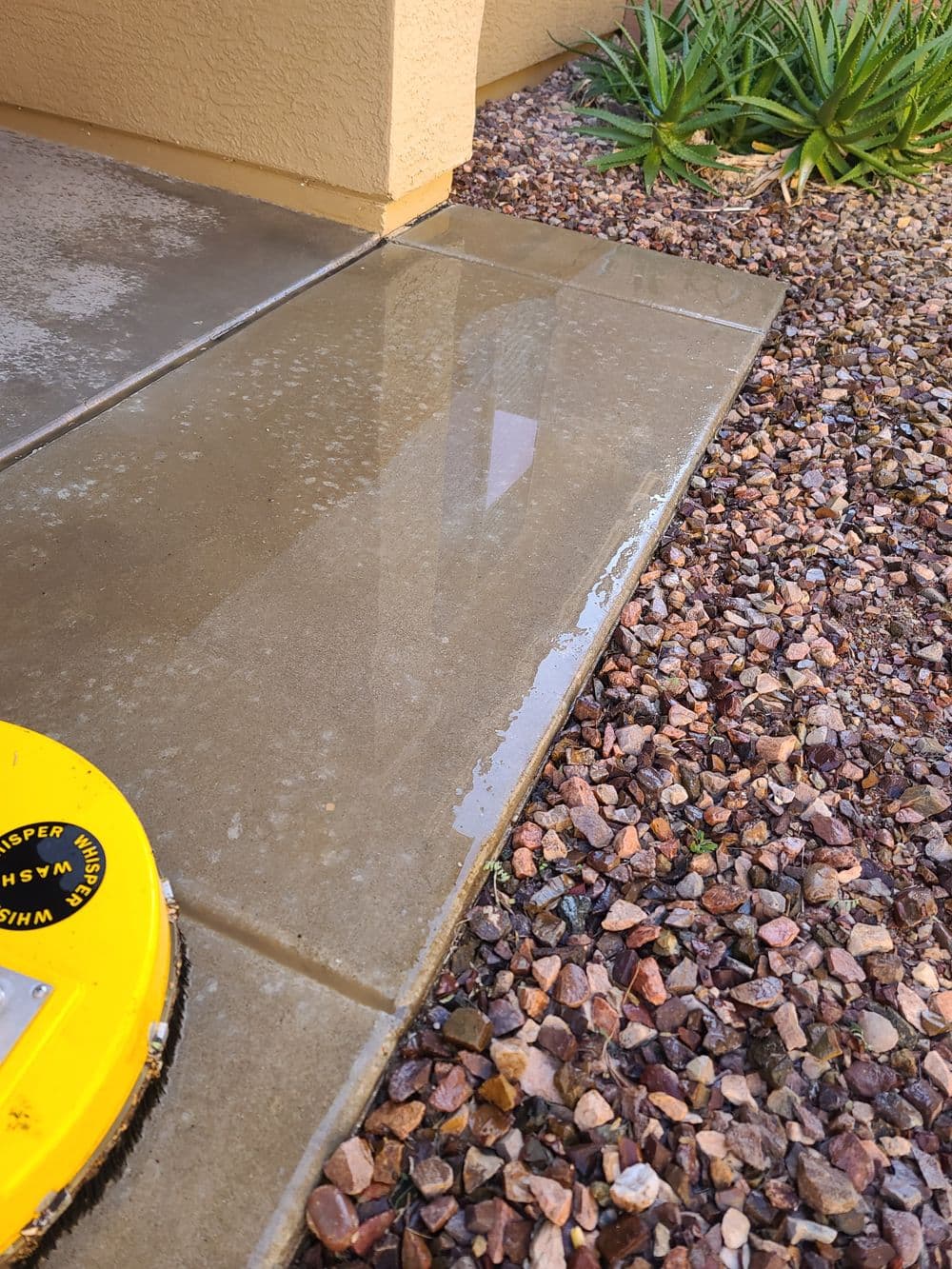 Wet concrete surface next to decorative gravel and a yellow utility cover.