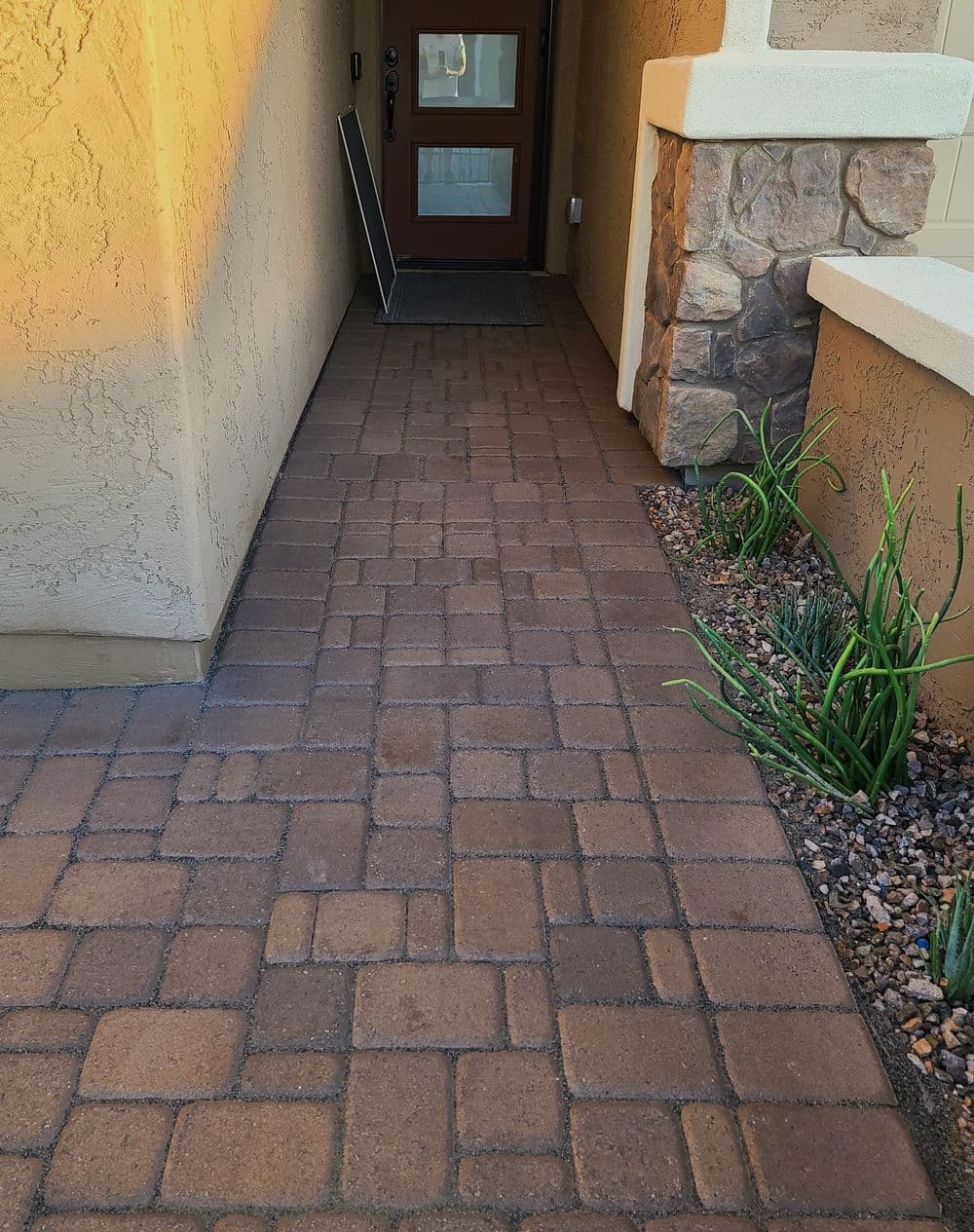 Paved pathway leading to a front door with stone accents and greenery along the sides.