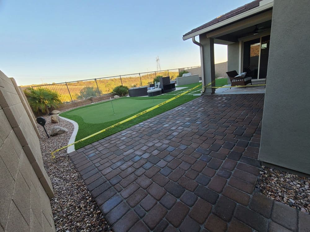 Backyard golf putting green with patio area and paver stones enclosed by a fence.