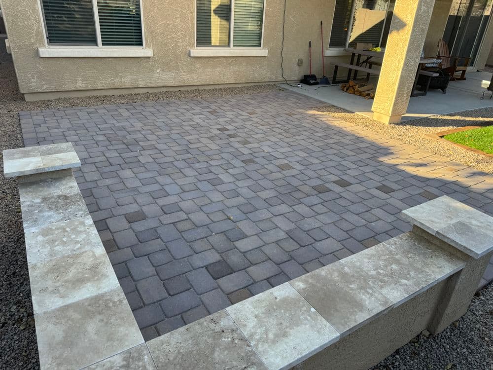 Paved outdoor patio with patterned stone and surrounding concrete border. Sunny residential setting.