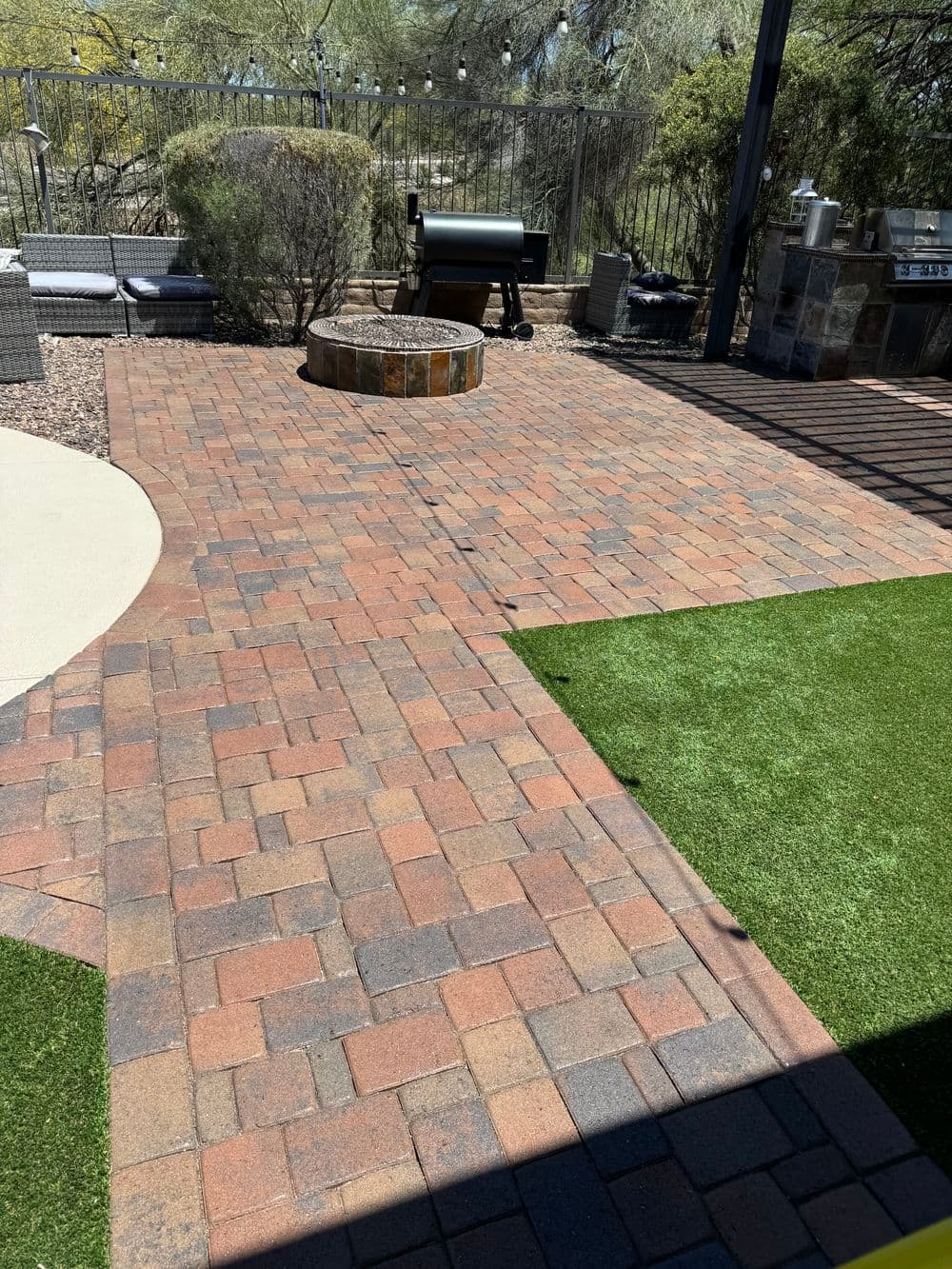 Paved patio with stone fire pit, barbecue grill, and artificial grass in a landscaped backyard.