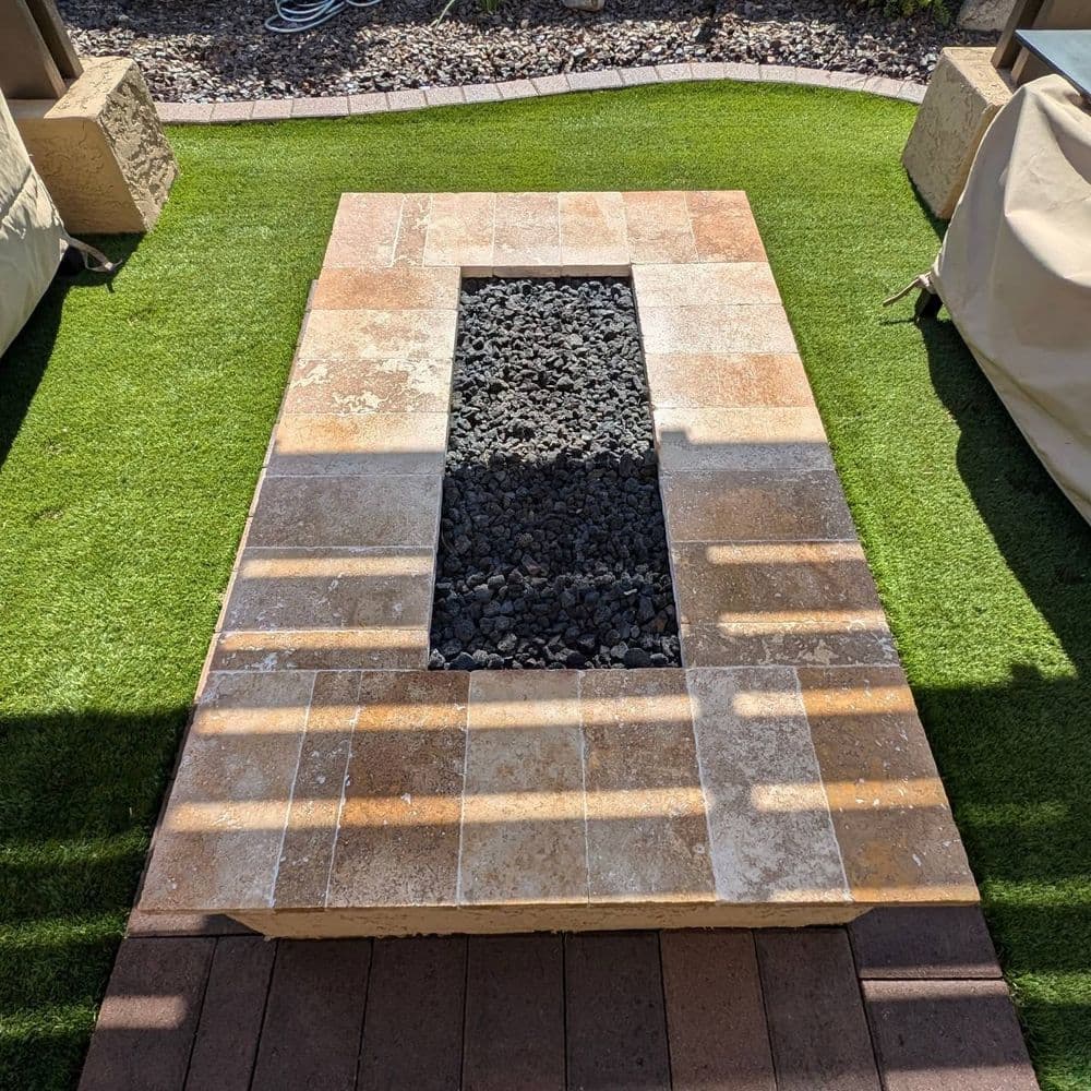 Rectangular outdoor fire pit with stone surround and black gravel, set on green artificial grass.