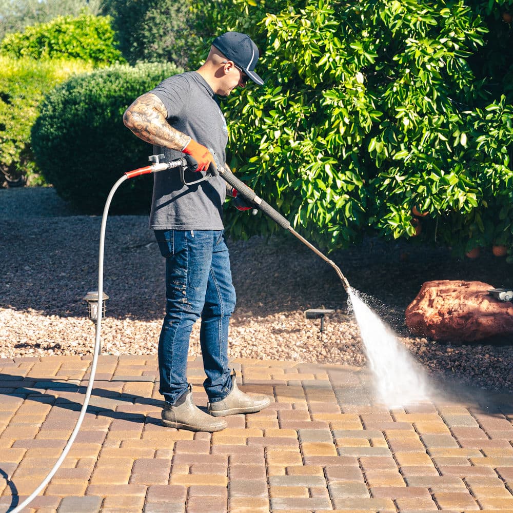 Man pressure washing brick patio in backyard, wearing gloves and cap on a sunny day.